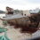 An old, damaged wooden boat is docked at a pier. The hull appears heavily deteriorated, with visible rot and missing planks. Green ropes are strewn around the dock.