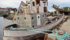 An old, dilapidated boat sits partially submerged and damaged at a dock, with debris and another damaged vessel nearby.