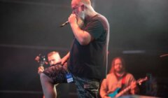 A man with a beard and microphone sings on stage, wearing a black shirt and camouflage shorts. Two other musicians are visible in the background performing.