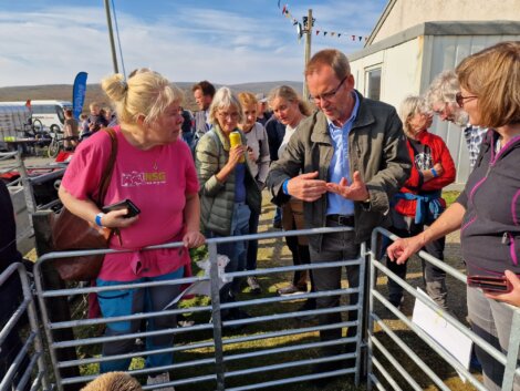 People gather around a metal enclosure outside. A man gestures with his hands while speaking to the group, which includes men and women dressed casually, appearing to listen attentively.