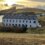 A large white building with multiple windows stands near the coast with surrounding grassy hills and cliffs under a cloudy sky with a setting sun.