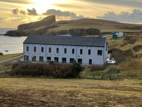 A large white building with multiple windows stands near the coast with surrounding grassy hills and cliffs under a cloudy sky with a setting sun.