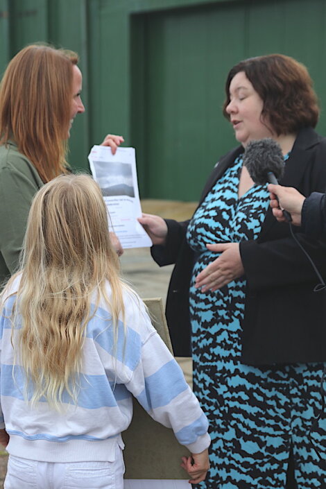 Two women and a child are standing together. One woman is holding a piece of paper while speaking into a microphone held by another person off-camera.