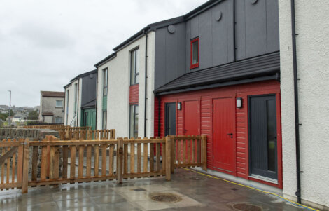 Row of modern, two-story terraced houses with wooden fences and paved front yards on a cloudy day.