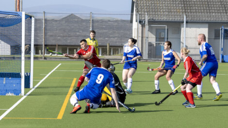 A field hockey game is in progress. Players in red and blue jerseys are near the goal, with some players attempting to score and others defending. The goalkeeper is on the ground. The umpire stands nearby.