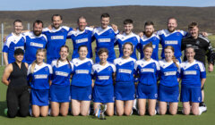 A sports team in blue and white uniforms poses on a field with a trophy, standing in two rows with a coach on the left and another individual on the right.