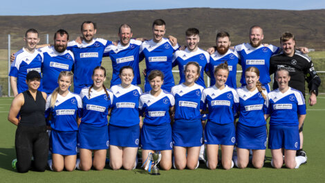 A sports team in blue and white uniforms poses on a field with a trophy, standing in two rows with a coach on the left and another individual on the right.