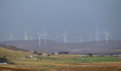 Wind turbines on a hill overlooking a rural landscape with scattered houses and green fields under a cloudy sky.