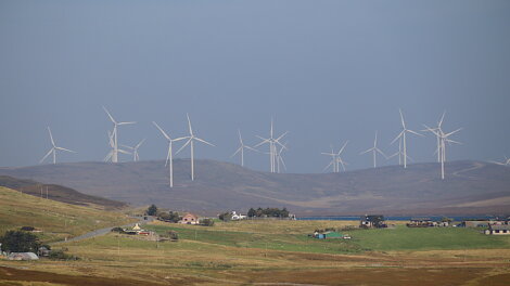 Wind turbines on a hill overlooking a rural landscape with scattered houses and green fields under a cloudy sky.