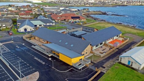 Aerial view of a coastal community showing a cluster of buildings near the shoreline, including residential homes and a modern building with a yellow accent, surrounded by parking lots and greenery.