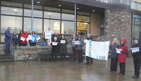 A group of people stands outside a building holding signs that read "Save Our Land" and other messages. It appears to be a protest or rally under rainy weather.