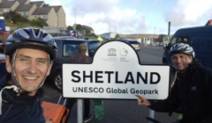 Two cyclists wearing helmets stand smiling by a sign reading "SHETLAND UNESCO Global Geopark," with parked vehicles and buildings visible in the background.