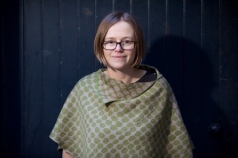 A person with glasses and short hair stands against a dark backdrop, wearing a green and gray patterned top.