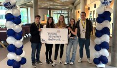 Five young individuals stand holding a banner that reads "Young Islanders Network" between two balloon columns with "Big Ideas" signs at the top in an indoor setting.