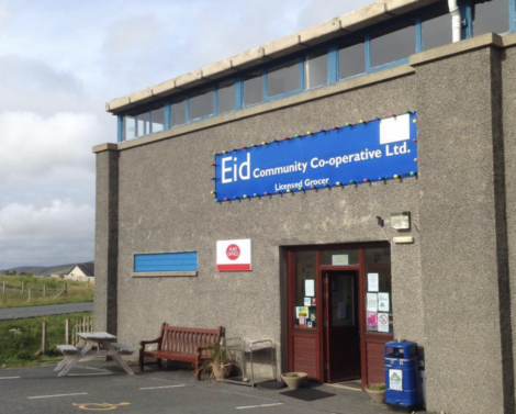 Storefront of Eid Community Co-operative Ltd. A bench and picnic table are outside. Several signs are displayed near the entrance, including one for a licensed grocer.