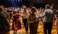 A group of people are dancing on a gymnasium floor under stage lights, engaging in what appears to be a social gathering or dance event.