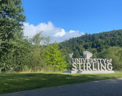 A large sign reading "University of Stirling" stands on a grassy area with trees and a hill in the background under a blue sky.