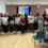 A group of people standing in a room holding certificates, with chairs and a presentation screen visible in the background.