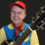 A person wearing a red cap, glasses, and a blue and yellow outfit is smiling and holding a guitar.