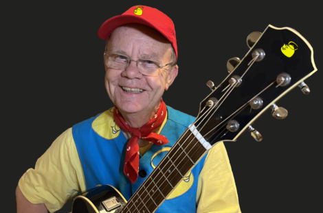 A person wearing a red cap, glasses, and a blue and yellow outfit is smiling and holding a guitar.