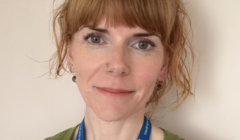 A person with auburn hair and a fringe is wearing a green cardigan and a blue lanyard. They are standing in front of a beige background.