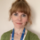 A person with auburn hair and a fringe is wearing a green cardigan and a blue lanyard. They are standing in front of a beige background.