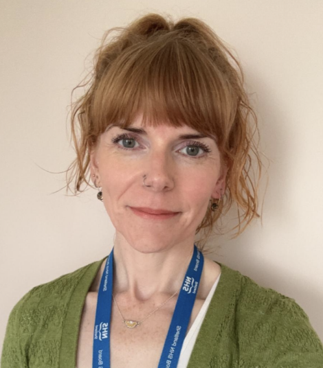 A person with auburn hair and a fringe is wearing a green cardigan and a blue lanyard. They are standing in front of a beige background.