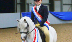 A person in a riding uniform and helmet smiles while seated on a white horse adorned with red, white, and blue ribbons in an indoor arena.