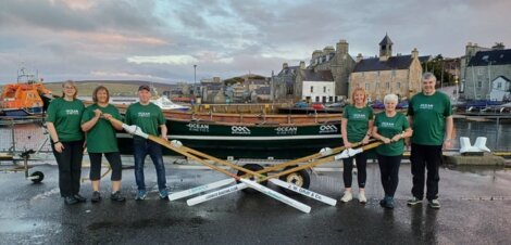 Group of six people wearing green shirts standing in front of a rowing boat on a trailer near a harbor with buildings and boats in the background.