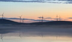 Landscape at dawn with several wind turbines situated on hills. Calm waters in the foreground reflect the turbines and the pastel-colored sky.