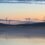 Landscape at dawn with several wind turbines situated on hills. Calm waters in the foreground reflect the turbines and the pastel-colored sky.