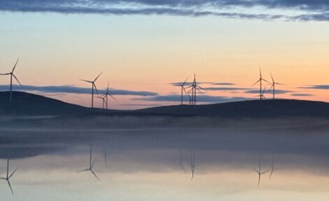 Landscape at dawn with several wind turbines situated on hills. Calm waters in the foreground reflect the turbines and the pastel-colored sky.