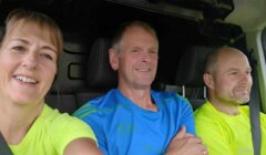 Three people in bright athletic shirts sitting in a car, smiling and looking ahead.