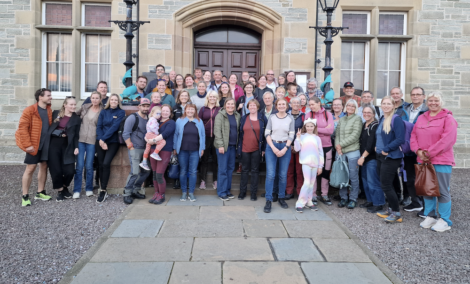 A large group of people of various ages posing together in front of a stone building with a wooden door.