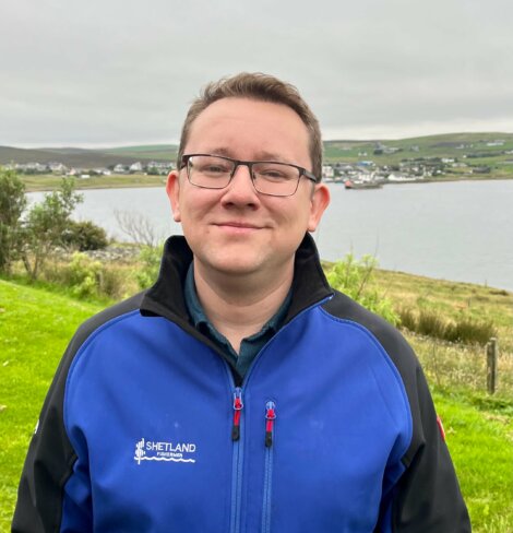 A person wearing glasses and a blue jacket stands outdoors with a body of water and a village in the background.