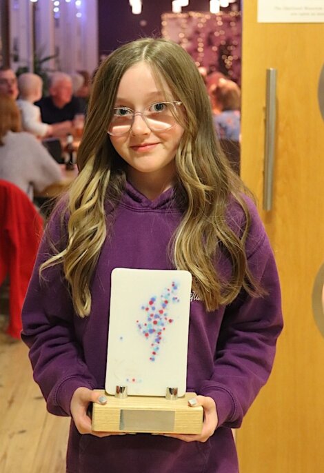 A young girl with long hair and glasses holds a rectangular award with a colorful dot design in front of a group of people in a room. She is wearing a purple sweater and standing near a wooden door.