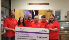 A group of six people in matching red shirts hold a large donation check for £227,538.69 to Cancer Research UK, standing inside Hay's Dock Café Restaurant.