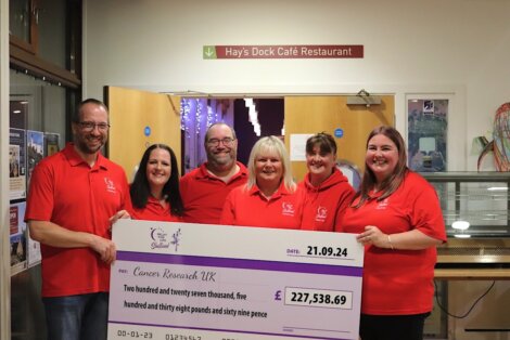 A group of six people in matching red shirts hold a large donation check for £227,538.69 to Cancer Research UK, standing inside Hay's Dock Café Restaurant.