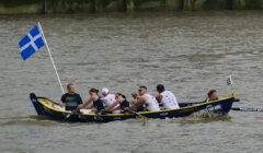 A rowing team of eight people is paddling in a boat with a blue and white flag on a river.