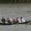 A rowing team of eight people is paddling in a boat with a blue and white flag on a river.