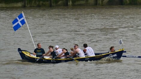 A rowing team of eight people is paddling in a boat with a blue and white flag on a river.