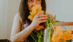 A person with long dark hair smelling a bouquet of yellow flowers while sitting on a bed beside a vase holding more flowers.