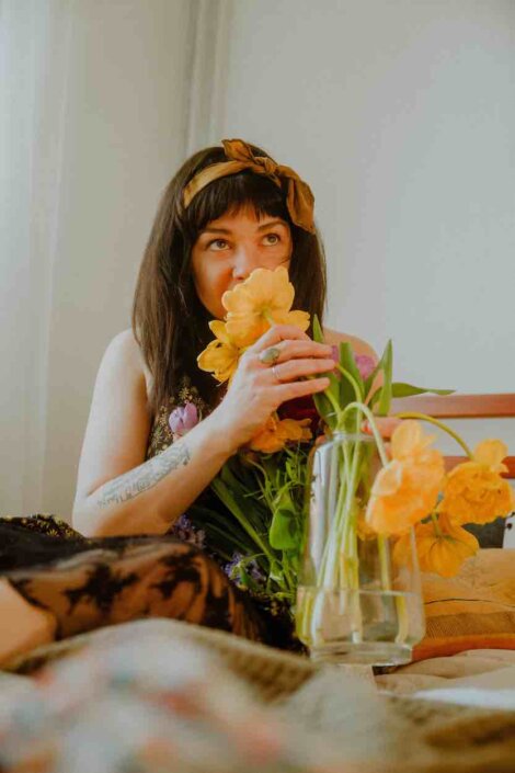 A person with long dark hair smelling a bouquet of yellow flowers while sitting on a bed beside a vase holding more flowers.