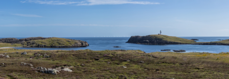 A panoramic view of a coastal landscape with grassy terrain, rocky hills, a lighthouse on an island, and several wind turbines visible on the horizon under a clear blue sky.