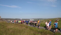 A group of runners on a countryside road participates in a race on a clear day, with fields and a distant ship in the background.