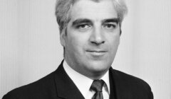 A black-and-white portrait of a man with gray hair wearing a dark suit and tie, looking at the camera with a neutral expression against a plain background.