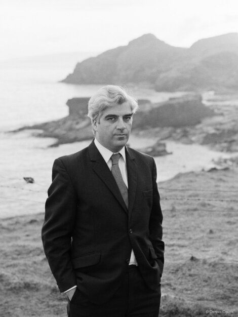 A man in a suit stands outdoors near a rocky coastline, looking at the camera.