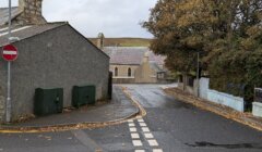 Lightly wet road with a no-entry sign, houses with stone walls, and autumn trees on both sides. A distant view of grassy fields under a cloudy sky in the background.