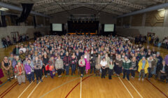 A large group of people standing in a gymnasium, facing the camera. In the background, there is a stage with a banner and a few people seated. Athletic markings are visible on the floor.