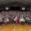 A large group of people standing in a gymnasium, facing the camera. In the background, there is a stage with a banner and a few people seated. Athletic markings are visible on the floor.